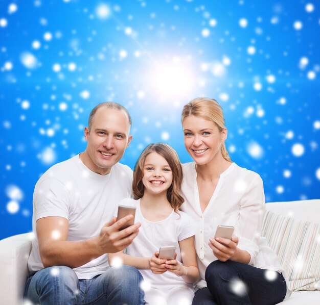 Foto familia, vacaciones de navidad, tecnología y concepto de personas - madre, padre y niña sonrientes con smartphones sobre fondo azul nevado