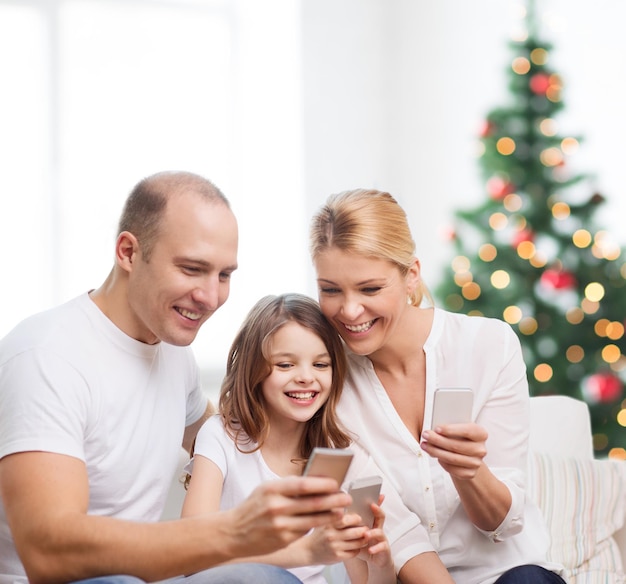 familia, vacaciones, tecnología y gente - madre, padre y niña sonrientes con smartphones sobre la sala de estar y el fondo del árbol de Navidad