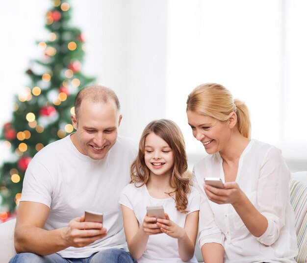 familia, vacaciones, tecnología y gente - madre, padre y niña sonrientes con smartphones sobre la sala de estar y el fondo del árbol de Navidad