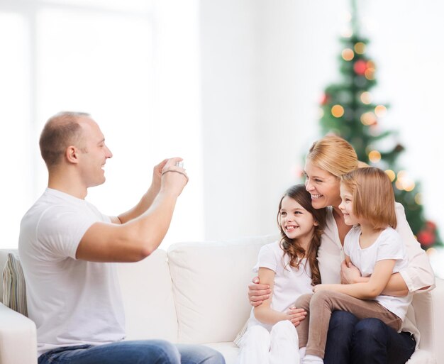 familia, vacaciones, tecnología y gente - madre, padre y niñas sonrientes con cámara sobre la sala de estar y el fondo del árbol de Navidad