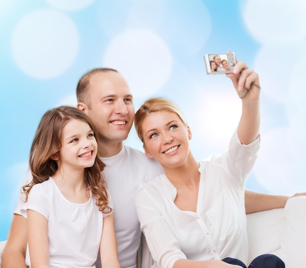 familia, vacaciones, tecnología y gente - madre sonriente, padre y niña haciendo selfie con cámara sobre fondo de luces azules