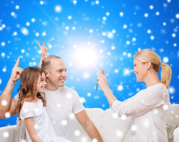 familia, vacaciones, tecnología navideña y concepto de personas: madre, padre y niña sonrientes con cámara sobre fondo azul nevado