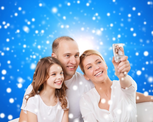 familia, vacaciones, tecnología navideña y concepto de personas: madre, padre y niña sonrientes haciendo selfie con cámara sobre fondo azul nevado