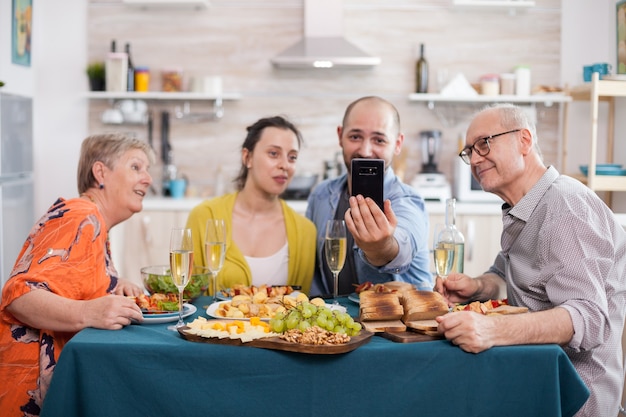 Familia de varias generaciones que usa el teléfono para videollamadas durante el brunch. Padres felices en la cocina vinculados con su hijo mayor y su esposa.