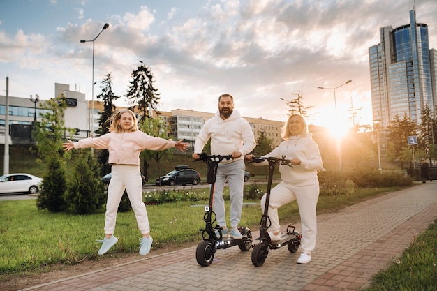 Una familia vestida de blanco se encuentra en la ciudad en patinetes eléctricos al atardecer.