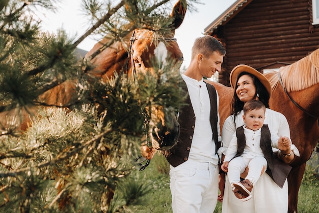 Una familia vestida de blanco con su hijo junto a dos hermosos caballos en la naturaleza. Una pareja elegante con un niño y caballos.