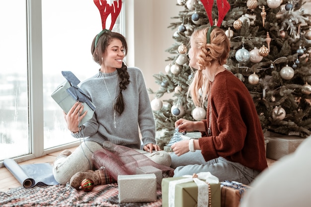 Familiares y amigos. Atractivas damas alegres que visten suéteres de gran tamaño y orejas de ciervo mientras se ocupan de la decoración de regalos Concepto de Navidad