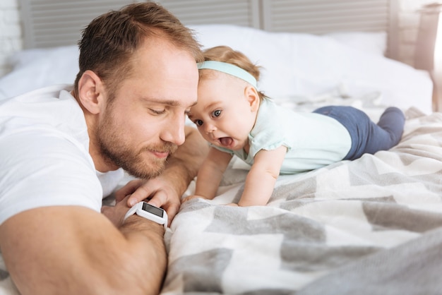 Familiarizar-se com as tecnologias modernas. Uma criança alegre e agradável deitada na cama com o pai e olhando para o smartwatch enquanto expressa interesse e alegria