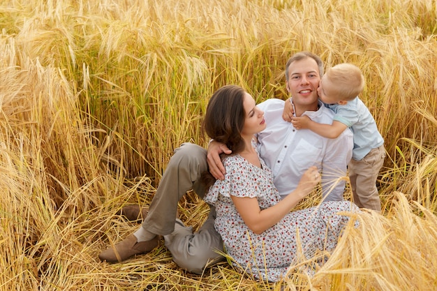 Familie, die auf dem Gras in einem Weizenfeld sitzt. Mutter, Vater und kleiner Sohn verbringen zusammen im Freien Freizeit. Eltern und Kind auf Sommerwiese