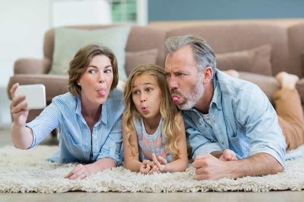 Familie, die selfie vom Handy nimmt, während sie zusammen auf Teppich im Wohnzimmer liegen