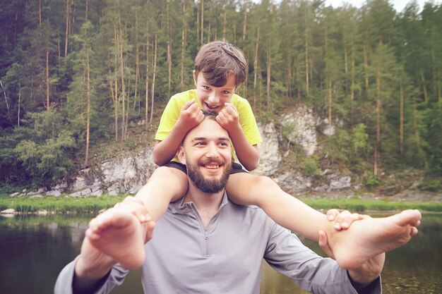 Familie geht durch Wald in der Nähe des Flusses, Vater trägt seinen Sohn auf den Schultern, Ökotourismus, Erholung im Freien während der Sommerferien.