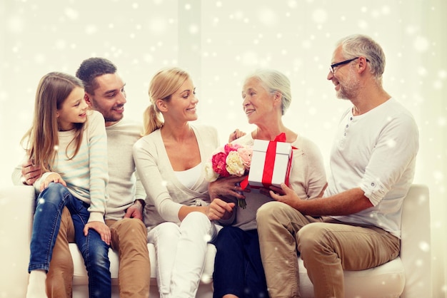 familie, glück, generation, urlaub und personenkonzept - glückliche familie mit blumenstrauß und geschenkbox, die zu hause auf der couch sitzt