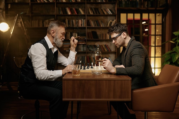 Familie intelligenter Menschen, die Schach spielen, Zigarren rauchen und Whiskey trinken. Ältere und jüngere Männer sitzen am Tisch und schauen auf das Schachbrett. Intelligente Spiele