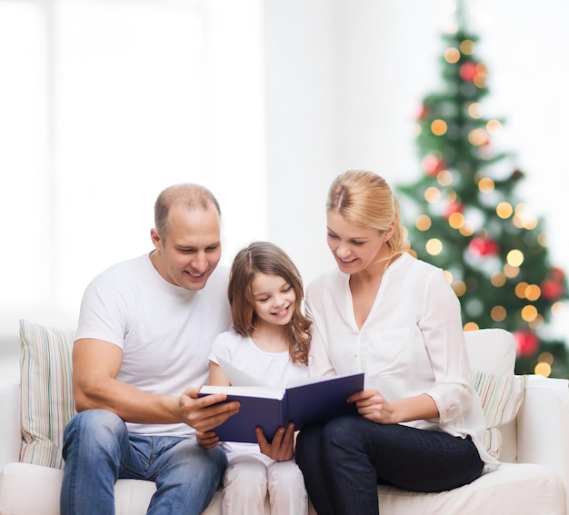 Familie, Kindheit, Feiertage und Menschen - lächelnde Mutter, Vater und kleines Mädchen lesen Buch über Wohnzimmer und Weihnachtsbaumhintergrund