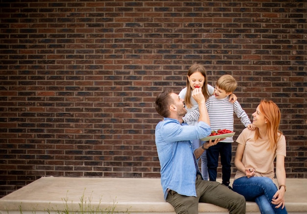 Familie mit Mutter, Vater, Sohn und Tochter, die draußen auf den Stufen der Veranda eines Backsteinhauses sitzen und Erdbeeren essen