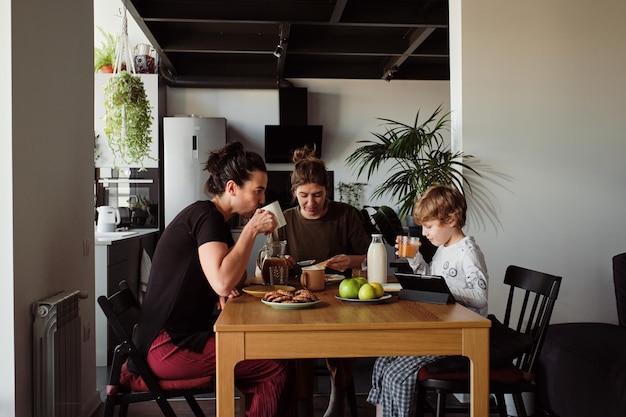 Familie mit zwei jungen Frauen mit kleinem Jungen, der am Tisch sitzt und in der Küche frühstückt