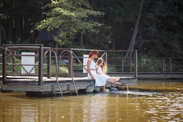 Familie, Mutter und Tochter sitzen am Fluss. Kleines Mädchen mit ihrer Mutter. Mädchen in weißen Kleidern. Familienzeit zusammen.