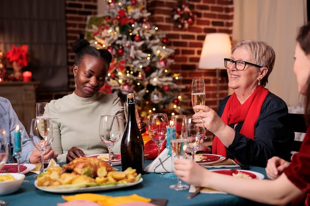 Familie trinkt Sekt, Frau mittleren Alters schlägt Weihnachtstoast beim festlichen Abendessen vor und hält Glas. Verschiedene Menschen feiern Winterferien und essen traditionelles Essen