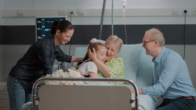 Familie überrascht Patientin im Ruhestand mit Blumen und Besuch auf der Krankenstation. Mutter und Kind besuchen kranke Oma im Bett, genießen Zeit mit Besuchern zur Erholung und Stärkung.