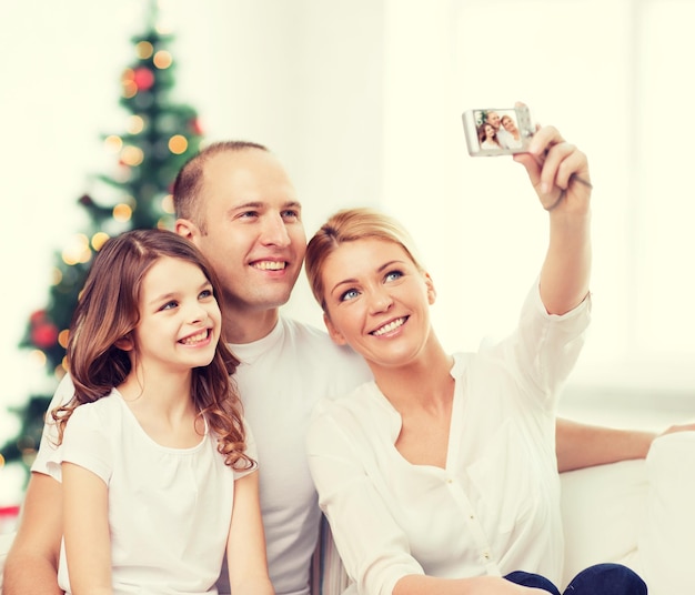 Familie, Urlaub, Technologie und Menschen - lächelnde Mutter, Vater und kleines Mädchen machen Selfie mit Kamera über Wohnzimmer und Weihnachtsbaumhintergrund