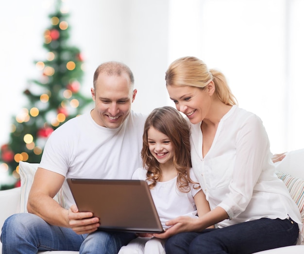 Familie, Urlaub, Technologie und Menschen - lächelnde Mutter, Vater und kleines Mädchen mit Laptop-Computer über Wohnzimmer und Weihnachtsbaumhintergrund