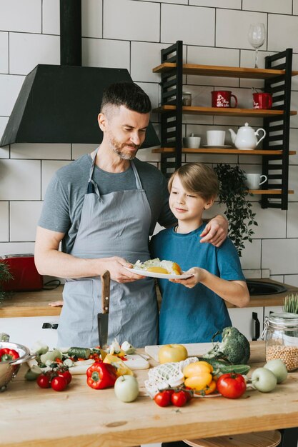 Familie, Vater, junger Mann und Sohn, Teenager, kochen Gemüsesalat in der Küche und verbringen schöne Zeit miteinander. Vater und Sohn unterhalten sich, kochen vegetarisches Essen und erledigen Hausarbeiten. 8. März und Muttertag