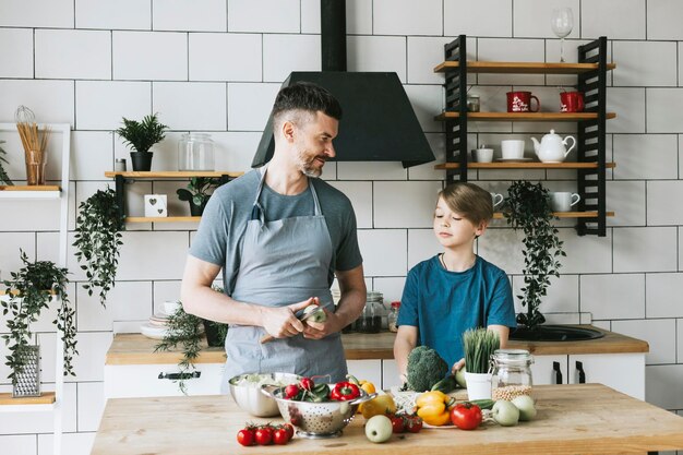 Familie, Vater, junger Mann und Sohn, Teenager, kochen Gemüsesalat in der Küche und verbringen schöne Zeit miteinander. Vater und Sohn unterhalten sich, kochen vegetarisches Essen und erledigen Hausarbeiten. 8. März und Muttertag