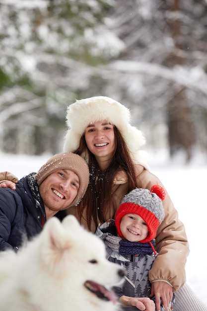 Familie von Mutter, Vater und Sohn, die sich im verschneiten Winterholz mit fröhlichem Schoßhund amüsieren