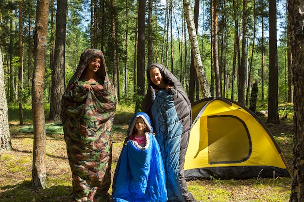 Familie von Touristen aus einem Vater, einer Mutter und einer kleinen Tochter posieren in Schlafsäcken in der Nähe eines Zeltes. Familienerholung im Freien, Inlandstourismus, Camping, Wanderausrüstung. Verpuppt wie Raupen-Humor