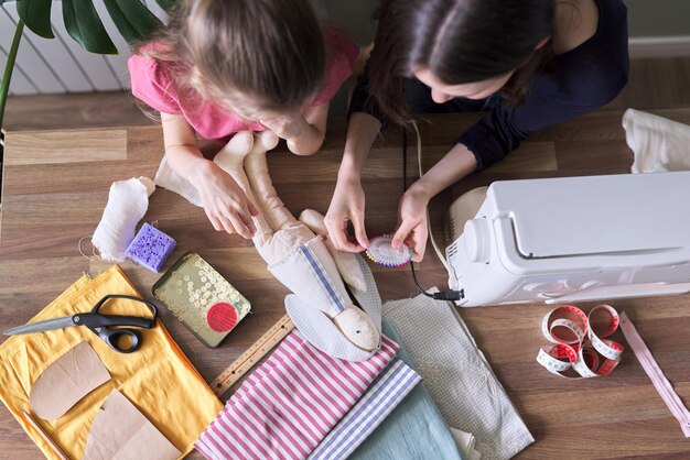 Foto familie, zwei mädchen, schwestern, teenager und jünger, die zusammen spielzeugpuppenhäschen nähen. hobbys, freizeit, kreativität und jugendliche fähigkeiten