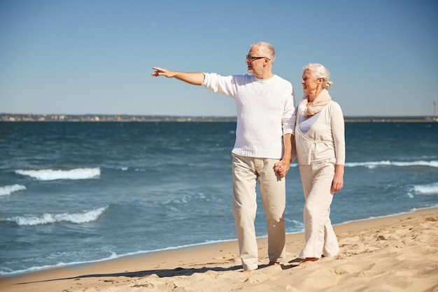 Familien-, Alters-, Reise-, Tourismus- und Personenkonzept - glückliches Seniorenpaar, das zu Fuß geht und mit dem Finger auf etwas am Sommerstrand zeigt