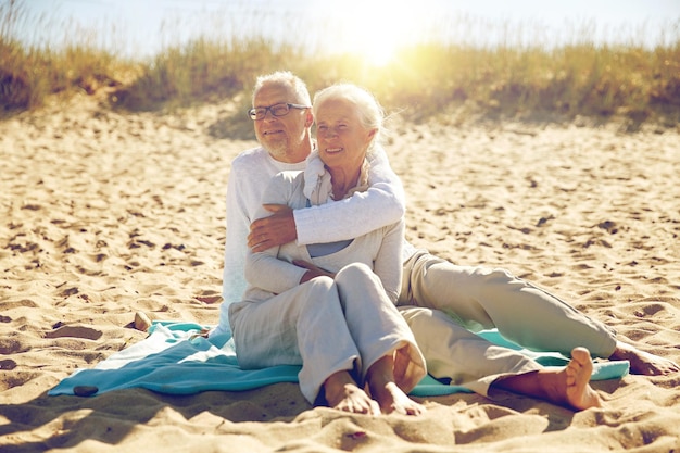 Familien-, Alters-, Reise-, Tourismus- und Personenkonzept - glückliches Seniorenpaar sitzt auf Plaid und umarmt sich am Sommerstrand