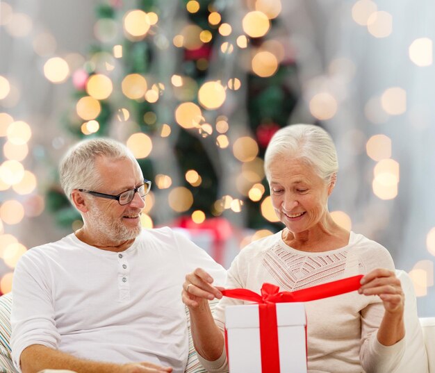 Familien-, Feiertags-, Alters- und Leutekonzept - glückliches älteres Paar mit Geschenkbox über Weihnachtsbaum beleuchtet Hintergrund