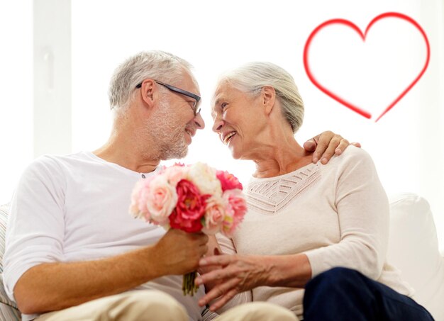 Familien-, Feiertags-, Alters- und Personenkonzept - glückliches Seniorenpaar, das zu Hause einen Blumenstrauß mit roter Herzform hält