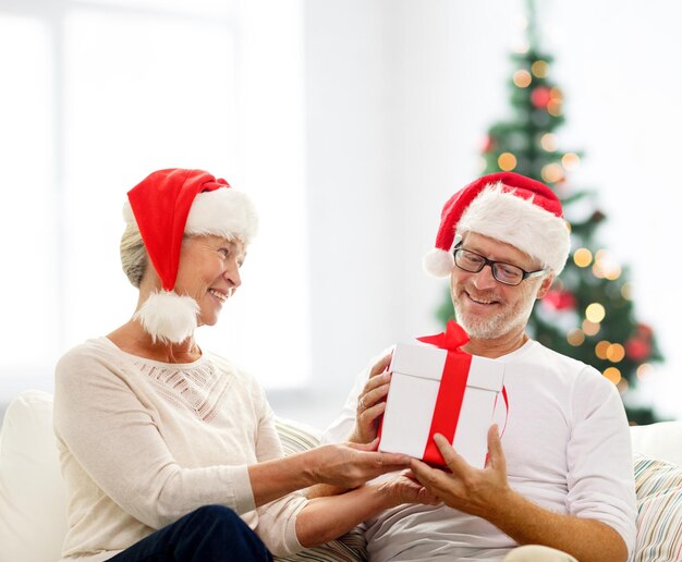 Familien-, Feiertags-, Alters- und Personenkonzept - glückliches Seniorenpaar in Weihnachtsmannmützen mit Geschenkbox über Wohnzimmer und Weihnachtsbaumhintergrund