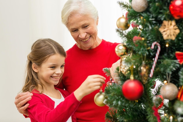 Familien-, Feiertags-, Generations- und Personenkonzept - lächelndes Mädchen mit Großmutter, die zu Hause Weihnachtsbaum schmückt