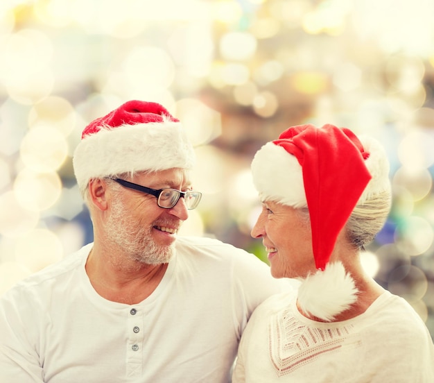 Familien-, Feiertags-, Weihnachts-, Alters- und Personenkonzept - glückliches Seniorenpaar in Weihnachtsmannmützen, das auf dem Sofa über hellem Hintergrund sitzt