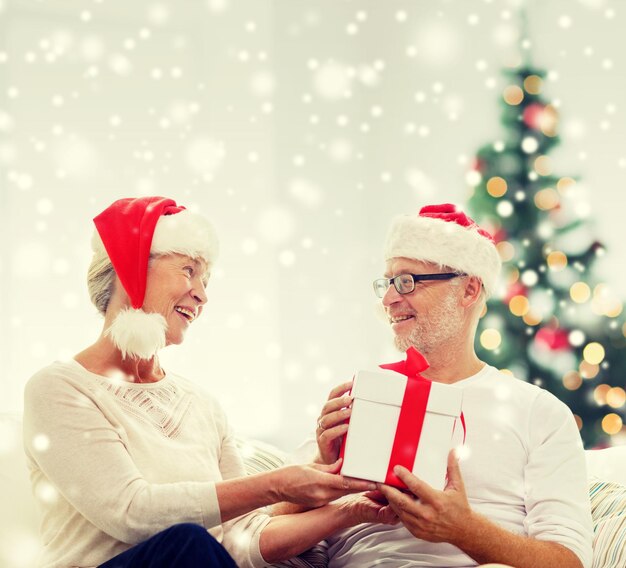 Familien-, Feiertags-, Weihnachts-, Alters- und Personenkonzept - glückliches Seniorenpaar in Weihnachtsmannmützen mit Geschenkbox zu Hause