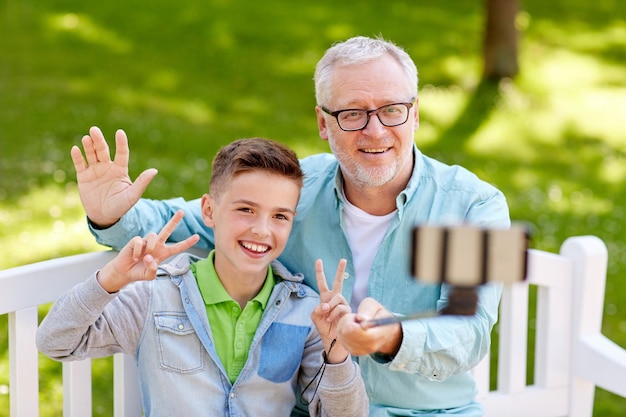 Familien-, Generations-, Technologie- und Menschenkonzept - glücklicher Großvater und Enkel fotografieren mit Smartphone-Selfie-Stick im Sommerpark