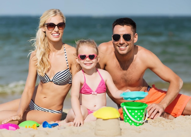 familien-, reise-, urlaubs- und personenkonzept - glücklicher mann, frau und kleines mädchen mit sonnenbrille spielen mit sandspielzeug am sommerstrand