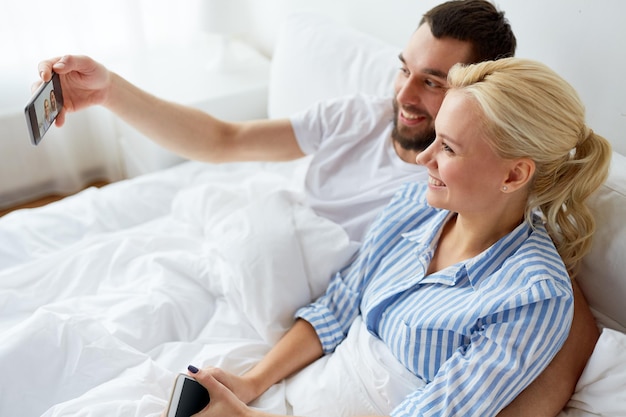 Familien-, Technologie- und Menschenkonzept - glückliches Paar mit Smartphone, das zu Hause ein Selfie im Bett macht