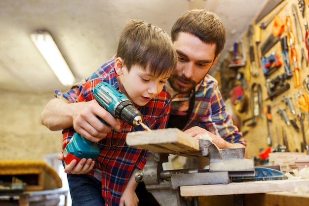 Familien-, Tischler-, Holzarbeiten- und Personenkonzept - Vater und kleiner Sohn mit Bohrer, der Holzbretter in der Werkstatt perforiert