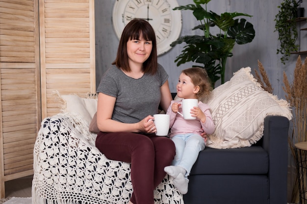 Familien- und Lifestyle-Konzept - junge Frau und ihre süße kleine Tochter sitzen und trinken Tee im Wohnzimmer