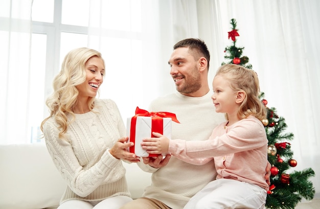 familien-, weihnachts-, feiertags- und menschenkonzept - glückliche mutter, vater und kleine tochter mit geschenkbox, die zu hause auf dem sofa sitzen