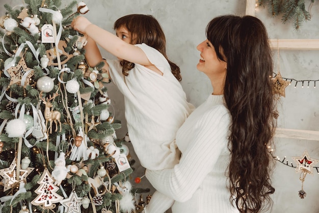 Familien-, Winterferien- und Menschenkonzept - glückliche Mutter hält kleine Tochter in den Armen und schmückt den Weihnachtsbaum zu Hause