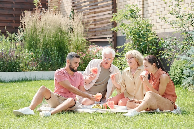 Familienaufnahme eines modernen älteren Mannes und einer Frau, die mit ihren erwachsenen Kindern auf dem Rasen picknicken