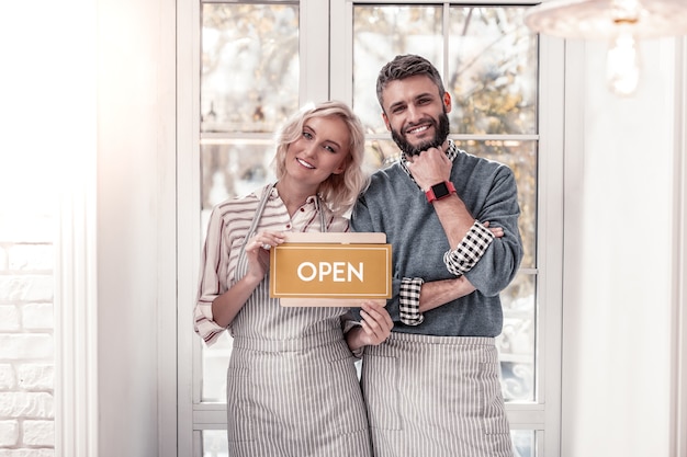 Familienbetrieb. Erfreutes fröhliches Paar, das zu Ihnen lächelt, während es sein eigenes Café eröffnet