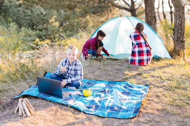 Familiencamping. Zwei Mädchen bauen ihr Zelt auf und ein Junge schaut sich die Show auf dem Laptop an