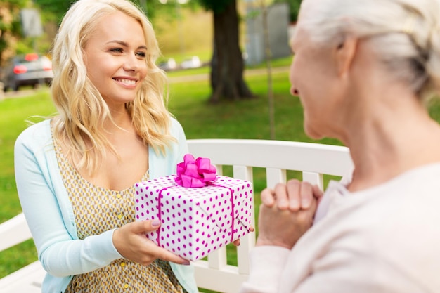 Familienfeiertage und Menschenkonzept glückliche lächelnde junge Tochter, die ihrer älteren Mutter ein Geschenk schenkt, sitzt auf einer Parkbank