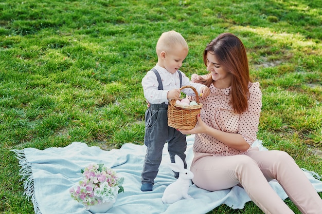 Familienfotosession der Mutter und des Babysohns für Ostern im Park, nahe bei ihnen ist ein Korb mit Eiern und einem Ostern-Kaninchen